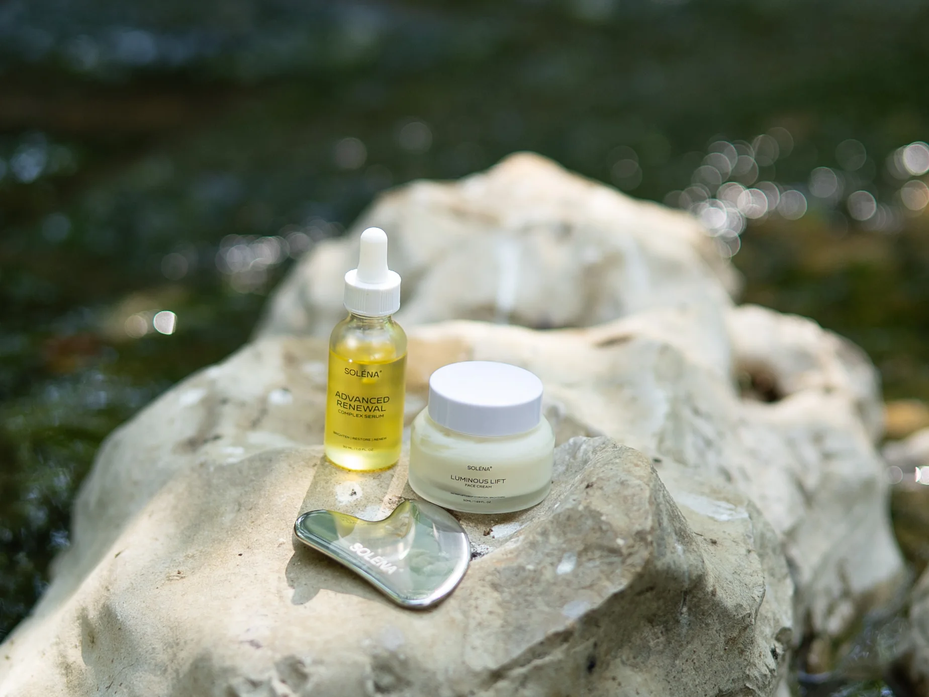 SOLÉNA serum, face cream and stainless steel Gua Sha beside a stream
